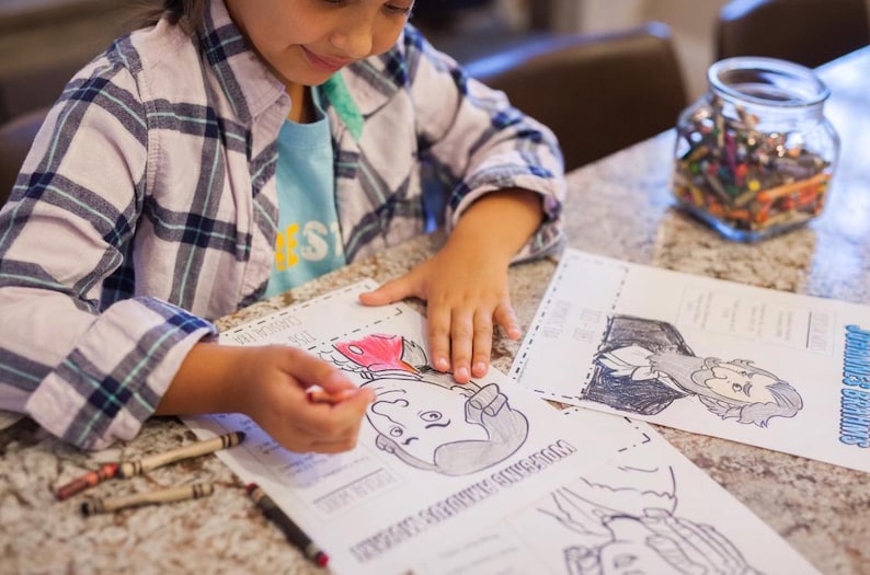 Puede incluir: Un ni&ntilde;o colorea un dibujo en blanco y negro de una figura hist&oacute;rica con un cray&oacute;n rojo. El ni&ntilde;o lleva una camisa a cuadros y una camiseta azul. Hay crayones y un tarro de crayones sobre la mesa.