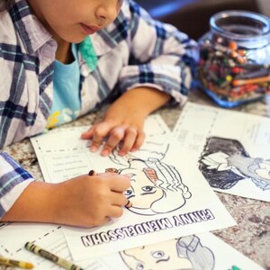 Puede incluir: Una persona colorea un dibujo en blanco y negro de una persona con el texto "FANNY MENDELSSOHN" en la p&aacute;gina. La persona lleva una camisa azul y una camisa a cuadros. Hay crayones y un tarro de crayones sobre la mesa.
