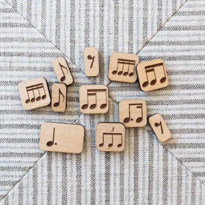May include: Wooden music note tiles with various symbols, including quarter notes, eighth notes, and a treble clef. The tiles are arranged on a striped fabric background.