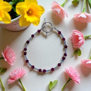 May include: A beaded bracelet with a silver toggle clasp. The bracelet features alternating dark red and light blue beads. The bracelet is displayed on a white surface, surrounded by pink flowers and yellow daffodils.