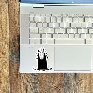 May include: A white laptop computer with a sticker of a black and white No Face character from the movie Spirited Away. The sticker has a white background and sparkles around the character. The text "I'm a little needy" is below the character.