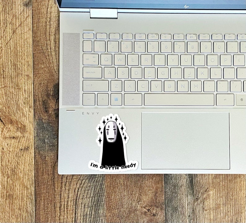 May include: A white laptop computer with a sticker of a black and white No Face character from the movie Spirited Away. The sticker has a white background and sparkles around the character. The text "I'm a little needy" is below the character.