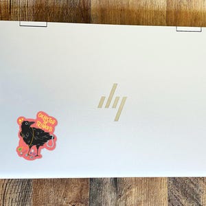 May include: A white laptop with a black crow sticker on the lid. The sticker says "Collector of Trinkets".