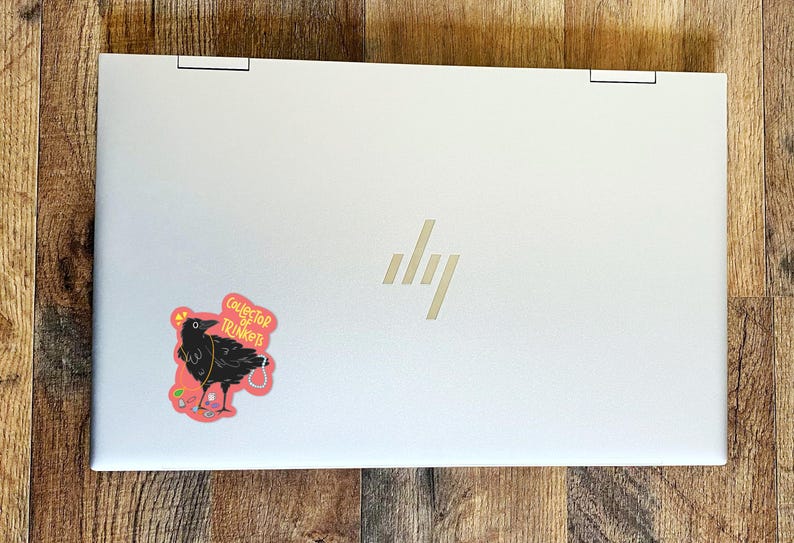 May include: A white laptop with a black crow sticker on the lid. The sticker says "Collector of Trinkets".