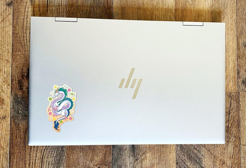May include: A silver laptop computer with a sticker of a purple and blue dragon with green accents on the lid. The dragon is surrounded by pink, yellow, and green flowers. The HP logo is visible on the lid.