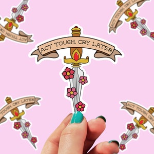 May include: A sticker with a gold dagger with a red gem and pink flowers. The dagger is surrounded by a banner with the text "ACT TOUGH, CRY LATER".