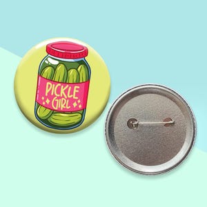 May include: A round button with a cartoon illustration of a jar of pickles. The jar is pink with the words "PICKLE GIRL" and a yellow background. The back of the button is silver.
