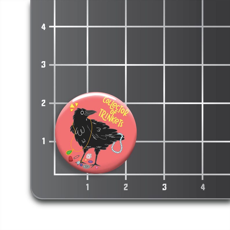 May include: A round, coral-colored button featuring a black crow adorned with jewelry. The crow is surrounded by trinkets and the text "Collector of Trinkets". The button is a fun accessory.