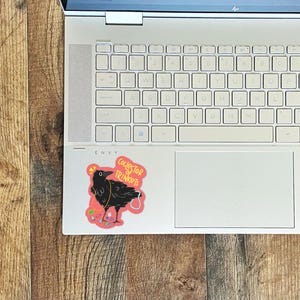 May include: A sticker of a black crow with a pink background. The crow is wearing a necklace of colorful trinkets and the text "Collector of Trinkets" is written in white on the sticker.