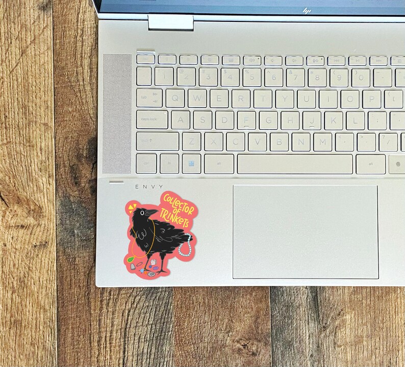 May include: A sticker of a black crow with a pink background. The crow is wearing a necklace of colorful trinkets and the text "Collector of Trinkets" is written in white on the sticker.