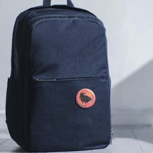 May include: A navy blue backpack with a small, round button pinned to the front. The button is red with a black crow illustration and the text "Crow Rukes" in yellow.