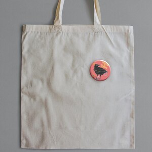 May include: A cream-colored tote bag with a round, pink button pinned to it. The button features a black crow illustration with the words "Angel Vibes" in a circular design. The crow is holding a small, decorated egg.