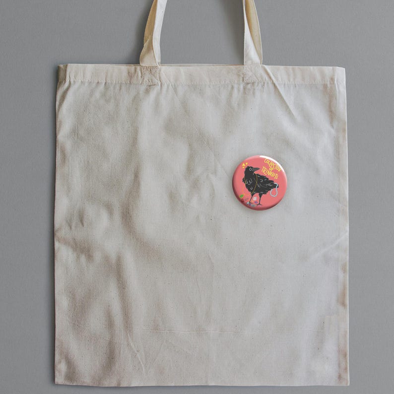 May include: A cream-colored tote bag with a round, pink button pinned to it. The button features a black crow illustration with the words "Angel Vibes" in a circular design. The crow is holding a small, decorated egg.