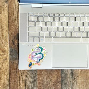 May include: A white laptop computer with a sticker of a blue and green dragon with pink and yellow accents on the lid. The sticker is on a white background with a light blue outline.
