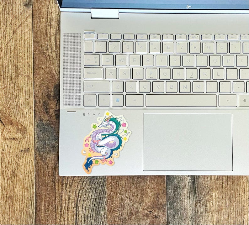 May include: A white laptop computer with a sticker of a blue and green dragon with pink and yellow accents on the lid. The sticker is on a white background with a light blue outline.