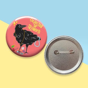 May include: A round, pink button with a cartoon raven illustration. The raven is black and holds a pearl necklace and other trinkets. Text on the button reads "Collector of Trinkets". The back of the button is silver.