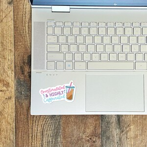 May include: A white laptop computer with a sticker on the top right corner. The sticker is pink, blue, and yellow and says "Overstimulated & HIGHLY Caffeinated" with a cartoon image of a cup of coffee with a straw.