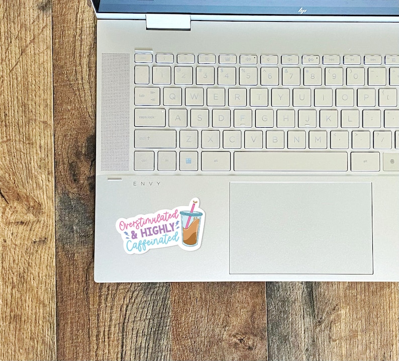 May include: A white laptop computer with a sticker on the top right corner. The sticker is pink, blue, and yellow and says "Overstimulated & HIGHLY Caffeinated" with a cartoon image of a cup of coffee with a straw.