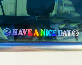 May include: A rainbow-colored decal applied to a car window featuring the text HAVE A NICE DAY in a bold, sans-serif font. The design is flanked by two smiling face icons, one on the far left and one on the far right, all in a gradient.