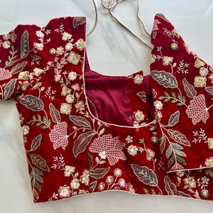 May include: A red velvet blouse with a floral embroidery design. The blouse has a scoop neck and short sleeves.