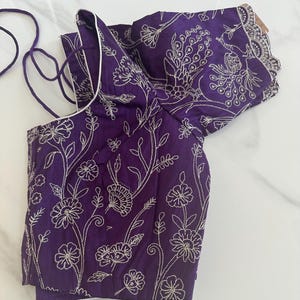 May include: A purple embroidered blouse with a white trim and a floral design. The blouse has a short sleeve and a tie closure at the neck.