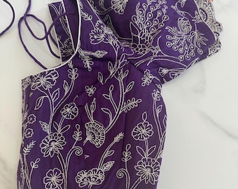 May include: A purple embroidered blouse with a white trim and a floral design. The blouse has a short sleeve and a tie closure at the neck.