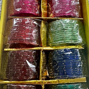 May include: A box of colorful bangles in shades of pink, red, green, and blue. Each set of bangles is wrapped in clear plastic. The bangles are decorated with small gold-colored embellishments.