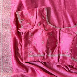May include: A vibrant pink saree and matching blouse. The saree features a detailed, embroidered border. The blouse has short sleeves and is adorned with delicate embroidery. The fabric has a subtle sheen, and the overall design is elegant.