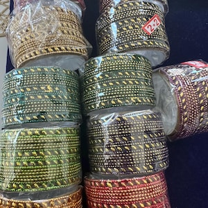 May include: An assortment of decorative trim rolls in various colors, including gold, green, red, and brown. Each roll is wrapped in clear plastic and features a repeating pattern of small, gold-colored embellishments. The trim appears to be made of a woven material.
