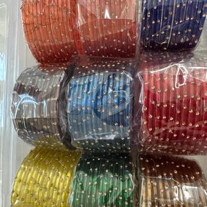 May include: A set of 12 colorful glass bangles with gold accents. The bangles are arranged in three rows of four, with each row featuring a different color. The colors include red, blue, brown, yellow, and green.
