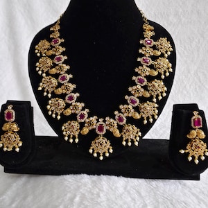 May include: A gold-toned necklace and matching earrings set displayed on a black stand. The necklace features a series of ornate gold elements, interspersed with rectangular red gemstones and small white pearls. The earrings mirror the necklace design.