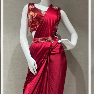 May include: A red satin saree with a floral print blouse and a beaded belt. The saree has a ruffled trim and is draped in a modern style.
