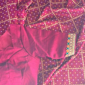 May include: A burgundy-colored blouse with a decorative sleeve cuff. The cuff features a detailed embroidered border with orange, green, and yellow accents. The fabric has a subtle sheen, and the background is a patterned textile.