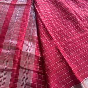 May include: A vibrant red fabric with a silver grid pattern and vertical stripes. The textile has a shimmering quality, with a detailed border at the bottom. The fabric is folded, showcasing its texture and design.