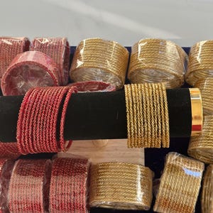 May include: A set of gold and red bangles. The bangles are stacked on a black stand and are made of a shiny, textured material.