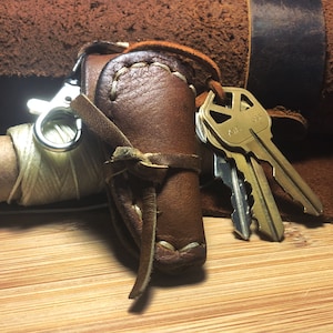 May include: A brown leather key holder with a loop for attaching keys. The holder has a small spool of thread attached to it. The holder is decorated with stitching and a braided leather strap. The holder is shown on a wooden surface with a set of keys attached to it.