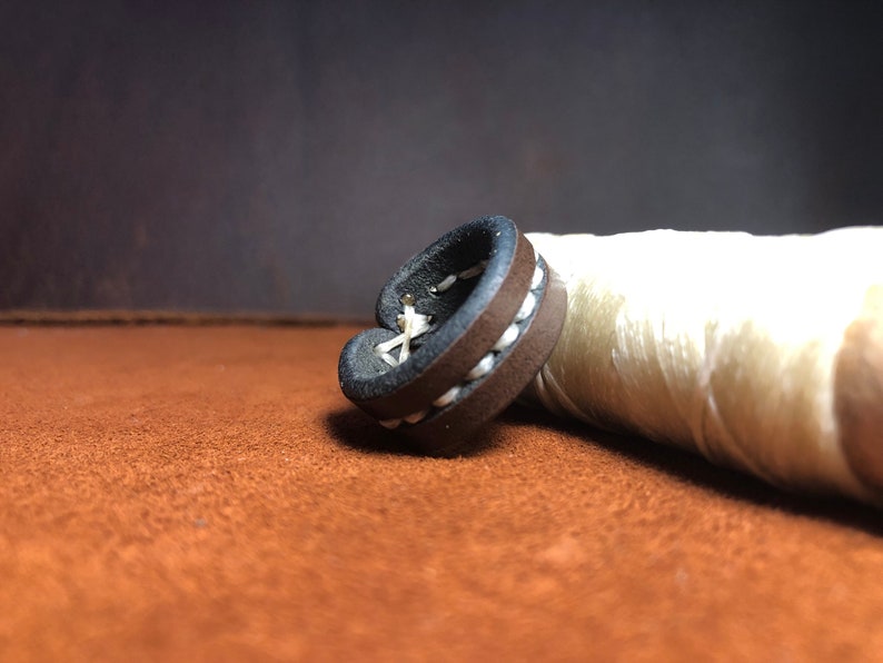 May include: A brown leather ring with white stitching. The ring is lying on a brown surface.