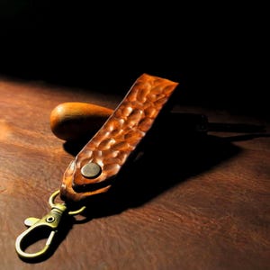 May include: A handcrafted leather keychain with a textured, hammered surface. The brown leather strap is attached to a brass-colored clasp. A wooden tool handle rests behind the strap, casting a shadow on the brown leather surface.