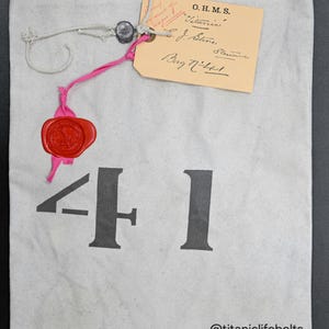 Puede incluir: Una bolsa de lona de color claro con el número "41" impreso en letras grandes de color gris oscuro. Una etiqueta beige con texto manuscrito está adjunta, junto con un sello de cera rojo. La bolsa está etiquetada "@titaniclifebelts replica."
