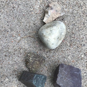 May include: A collection of four rocks of different shapes, sizes, and colors. The rocks are on a gray concrete surface. The rocks are a light gray, dark gray, brown, and reddish brown.