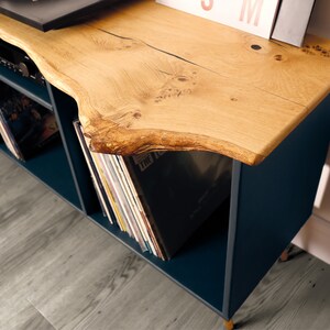Może przedstawiać: Stojak na gramofon w kolorze ciemnoniebieskim z blatem z żywego drewna. Stojak ma dwie p&oacute;łki do przechowywania płyt. Stojak stoi na jasnoszarym drewnianym podłodze.