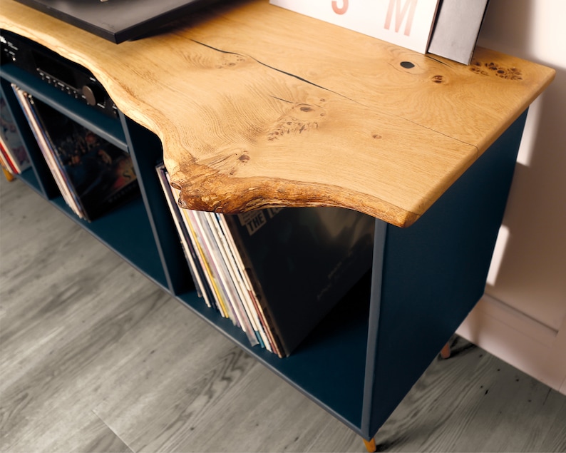 Może przedstawiać: Stojak na gramofon w kolorze ciemnoniebieskim z blatem z żywego drewna. Stojak ma dwie p&oacute;łki do przechowywania płyt. Stojak stoi na jasnoszarym drewnianym podłodze.