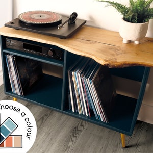 May include: A dark blue record player stand with a live edge wood top. The stand has two shelves for storing records and a space for a stereo receiver. The stand has gold legs and is sitting on a wood floor.