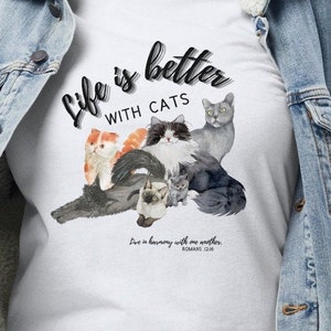 May include: White t-shirt featuring a watercolor illustration of several cats in various colors and patterns. The text "Life is better with cats" is written above the cats in a script font. Below the cats is the text "Live in harmony with one another. Romans 12:16."