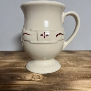 May include: A white ceramic mug with a red floral design and a footed base. The mug has a handle and is decorated with a red floral pattern.