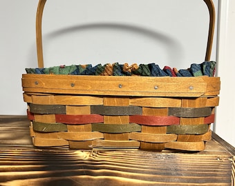 Könnte beinhalten: Ein geflochtener Korb mit Holzgriff. Der Korb hat eine Mischung aus Farben, darunter Rot, Grün und Braun. Der obere Rand ist mit einem gemusterten Stoff in Blau-, Grün- und Brauntönen gefüttert.