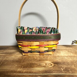May include: A small, woven basket with a wooden handle. The basket is lined with a floral fabric and has a green, purple, and yellow trim.