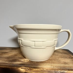 May include: A white ceramic mixing bowl with a woven design and a handle. The bowl is sitting on a wooden surface.