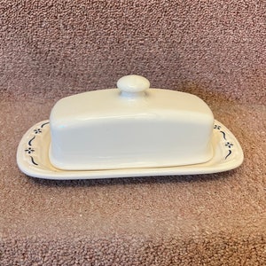 May include: A white ceramic butter dish with a blue floral design around the edge of the base. The lid has a knob on top.