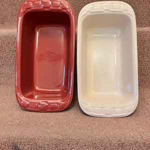 May include: Two rectangular ceramic loaf pans. One is a deep red color, and the other is off-white. Both pans have a glossy finish and a decorative woven pattern around the rim. They are empty and ready for baking.